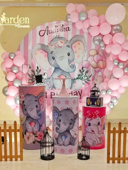 A sweet elephant-themed party in pink and grey. The setup includes a large circular backdrop, themed plinths, and a cute wooden fence.