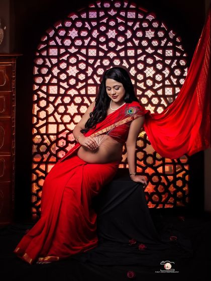 In this striking portrait, a mom-to-be is seated in a vibrant red saree, her bare bump adorned with a delicate waist chain. The intricate 'jaali' screen behind her creates a beautiful play of light and shadow.