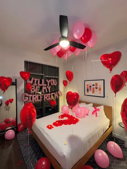 I can help you ask the big question with a beautiful surprise setup. This room features 'WILL YOU BE MY GIRLFRIEND' foil balloons, a mix of red and pink latex and heart-shaped foil balloons, and a bed covered in rose petals to create the perfect romantic proposal setting.