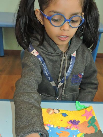 A student with glasses adds the final touches to her vibrant and detailed clay diorama of a beach scene.