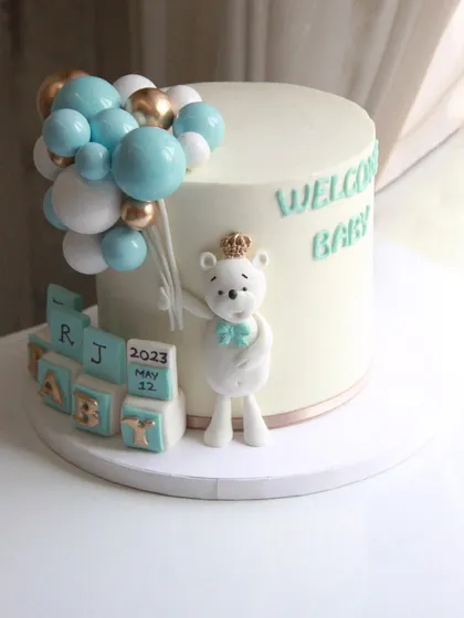 A precious welcome baby cake in shades of blue and white, featuring a cute teddy bear holding balloons and personalized blocks with the baby's initials and birth date.