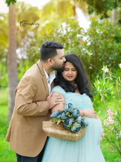 A sweet couple's portrait in a garden, with the mother-to-be in a light blue gown holding a basket of flowers.
