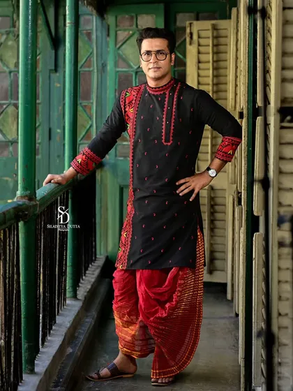A full-length shot of Anirban Bhattacharya, showcasing his complete traditional outfit with red dhoti pants. The composition emphasizes the architectural details of the old Kolkata balcony.