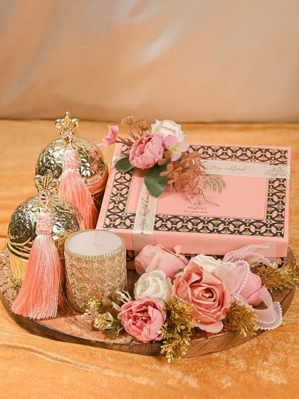 A truly royal Rakhi hamper. This arrangement on a wooden tray features a decorative box, a scented candle, and ornate golden spheres with tassels, all surrounded by beautiful pink flowers.