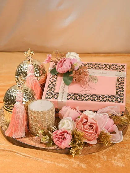 A truly royal Rakhi hamper. This arrangement on a wooden tray features a decorative box, a scented candle, and ornate golden spheres with tassels, all surrounded by beautiful pink flowers.