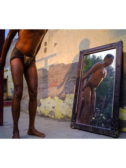 A wrestler looks at his reflection in a mirror propped against a wall at an outdoor gym. The golden hour light and the subject's pose create a moment of introspection and pride.