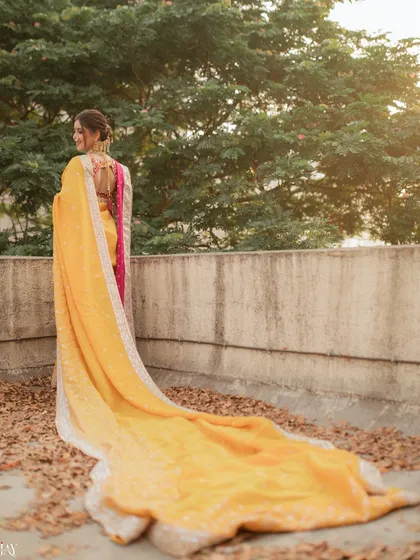 This shot from behind showcases the dramatic and beautiful trail of the bride's yellow nauvari saree against a bed of autumn leaves. It highlights the elegance and grandeur of the traditional Maharashtrian bridal look.
