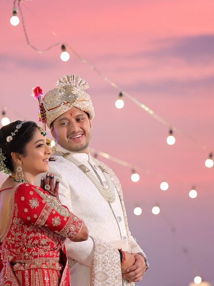 A romantic shot of the couple against a beautiful pink and purple sunset sky. The string lights add a magical touch to this candid wedding portrait.