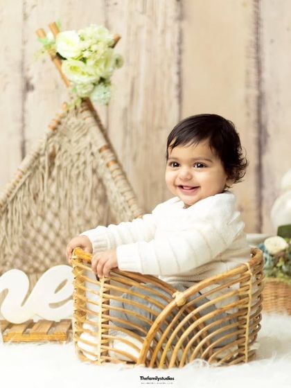 A happy baby boy in a basket during his first birthday photoshoot, featuring a rustic theme with a teddy bear and 'One' sign.