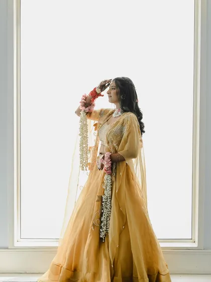 A high-key portrait against a white background, creating a soft and ethereal look. The bride, holding her floral kaleeras, looks like a vision in her golden-yellow lehenga.
