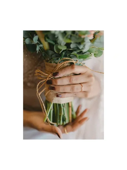 A close-up of the bride's hands holding her rustic bouquet. The image focuses on her simple, elegant ring and the natural texture of the burlap-wrapped stems.