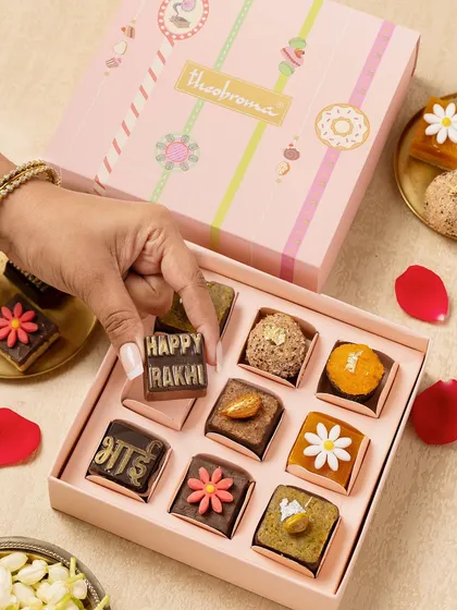 This shot captures the essence of a festive Rakhi celebration. The hand placing the "Happy Rakhi" chocolate adds a personal touch, while the surrounding props like rose petals and jasmine create a rich, cultural context.