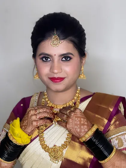 Adjusting the final details for this bride's Muhurtham look. The focus is on traditional gold jewellery and a classic red lip that complements her off-white and maroon silk saree.