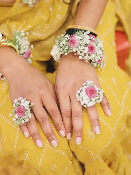 A close-up of the bride's hands, showing the detailed hath phool and floral rings. The design combines pink flowers with baby's breath and pearl accents, matching the rest of her Haldi jewellery.