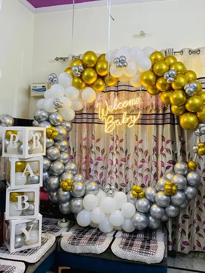 A 'Welcome Baby' decoration for an intimate home setting. This design uses a full ring of silver, gold, and white balloons, a neon sign, and 'BABY' blocks.