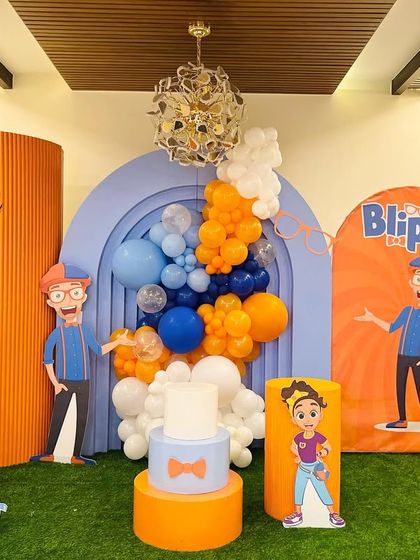 A full view of the Blippi birthday party for Ubadah. The bright orange and blue setup includes multiple character cutouts, themed backdrops, and a large light-up number '7'.