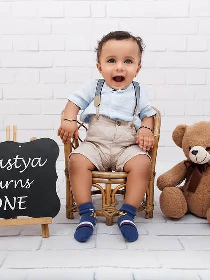 "Agastya turns ONE." A classic first birthday portrait with a chalkboard sign and a friendly teddy bear.