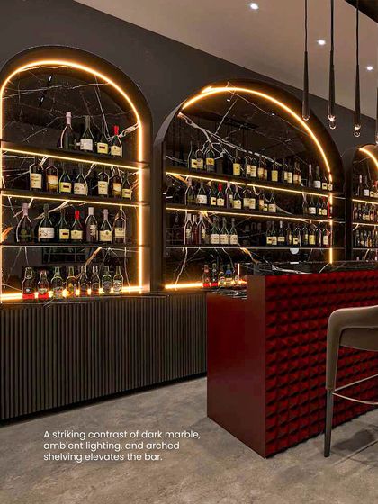 The bar is elevated by a striking contrast of dark marble, warm ambient lighting, and arched shelving. The red, tufted bar front adds a pop of color and texture.