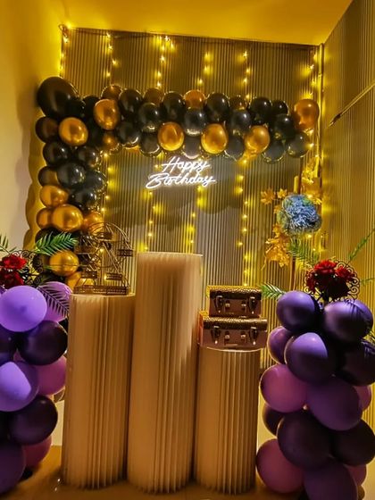 An elegant birthday setup with a black and gold balloon arch, accented with purple balloon columns. The decor includes a mini Ferris wheel and floral arrangements.