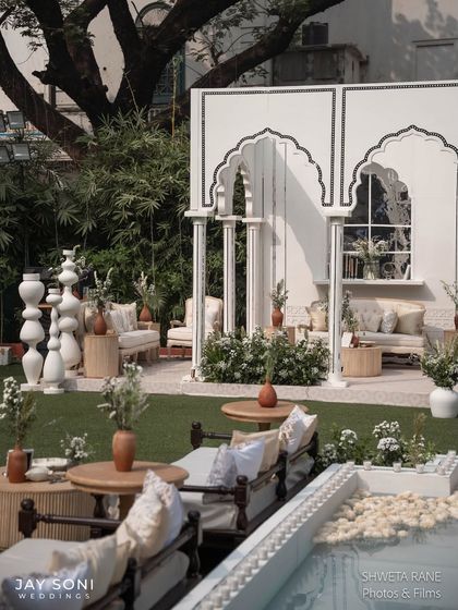 An outdoor lounge area designed with a clean, monochromatic palette. The white architectural backdrop with traditional arches complements the mix of modern and classic ivory seating.