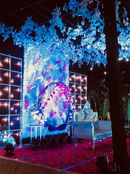 A dramatic stage design featuring a full wall of white flowers with pink accents, illuminated by cool blue lighting. The artificial tree with blue leaves adds a unique, artistic touch to the entire setup.