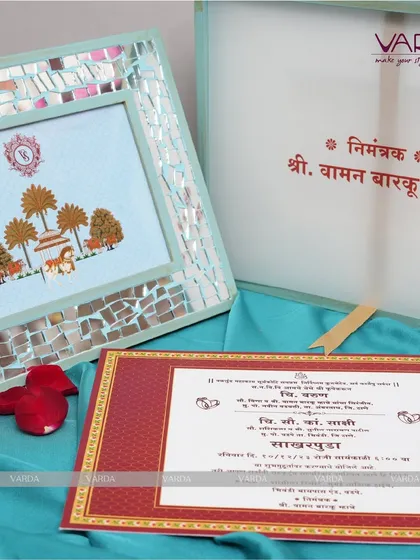 A unique invitation concept featuring a framed artwork with a traditional Indian motif and a mirrored border. This reusable frame is paired with a classic red and gold invitation card.