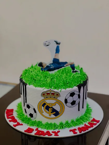 A fantastic Real Madrid football cake for a fan's birthday. The cake features a grass-like green topping, the team logo, and a player doing a bicycle kick.