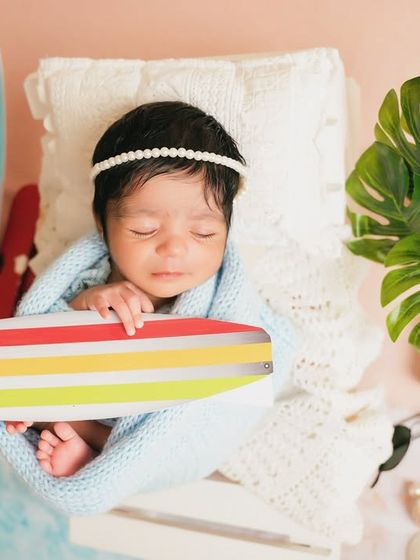 A newborn swaddled in blue holds a tiny surfboard, part of a creative beach-themed setup. It's a playful way to dream of future family holidays.