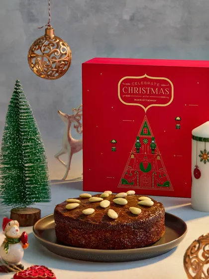This image features a classic Christmas almond cake alongside its beautiful red gift box from Marriott Bonvoy. The composition with miniature trees and ornaments creates a complete and charming holiday gifting story.