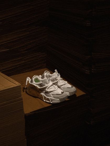 A dark and moody shot of white sneakers placed in a cardboard structure. The dramatic shadows and unique setting create an urban, industrial vibe for this streetwear brand.