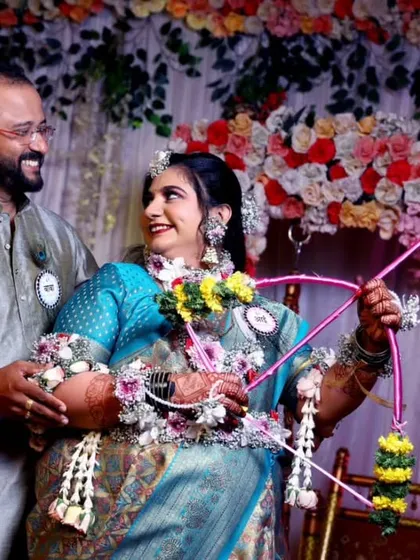 A happy couple enjoying their baby shower. The mom-to-be is adorned with floral jewelry and holds the traditional bow and arrow prop in front of a 'Kunitari yenar ga' sign, capturing a perfect moment.