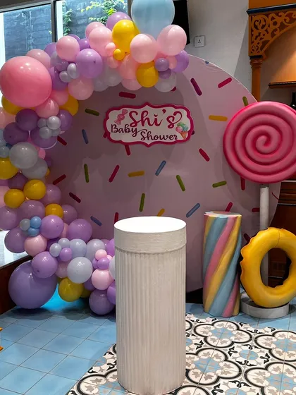 A sweet and playful "Candyland" theme for a baby shower. The backdrop looks like a giant donut with sprinkles, framed by a pastel balloon arch and oversized candy props like a lollipop and marshmallow twist.