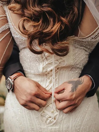 A detail shot of the groom's hands lacing up the bride's corseted gown, an intimate and symbolic moment of togetherness.