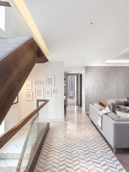The AMARA residence features a beautiful wooden staircase that ascends alongside a chevron-patterned marble floor. The living area is decorated in soothing grey tones with a textured feature wall, creating a harmonious and elegant interior.