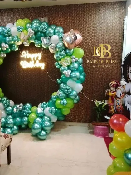 A modern Janmashtami celebration setup featuring a balloon ring in shades of green and silver, a 'Happy Birthday' neon sign, and a large cutout of little Krishna.