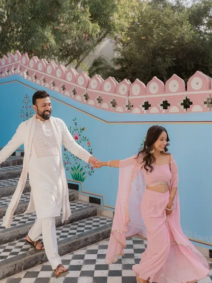 A stylish couple at their Haldi event in Rajasthan. Their coordinated pink and white outfits pop against the colorful blue and pink walls of the heritage venue.