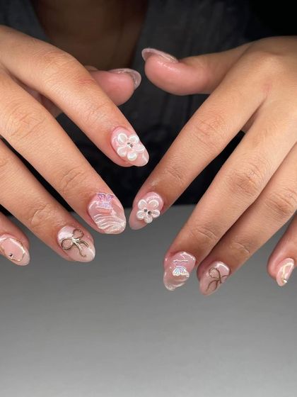 A delicate and ethereal design with 3D white flowers, gold bow charms, and a subtle shimmer base. This is a beautiful example of soft, romantic nail art.