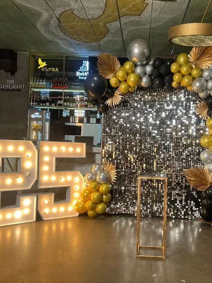 For a 25th anniversary party, I created this glamorous photo corner. It includes a shimmering silver sequin backdrop, large light-up '25' marquee numbers, and a custom balloon arch in a sophisticated black, gold, and silver color scheme.
