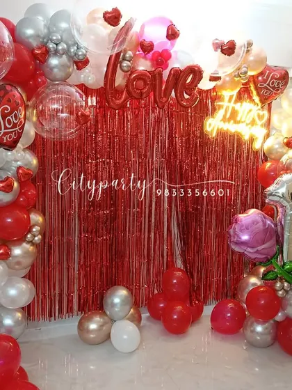 We designed this vibrant 10th-anniversary decoration with a red tinsel backdrop, a 'Happy Anniversary' neon sign, and a balloon arch in red, silver, and rose gold for a romantic celebration at home.