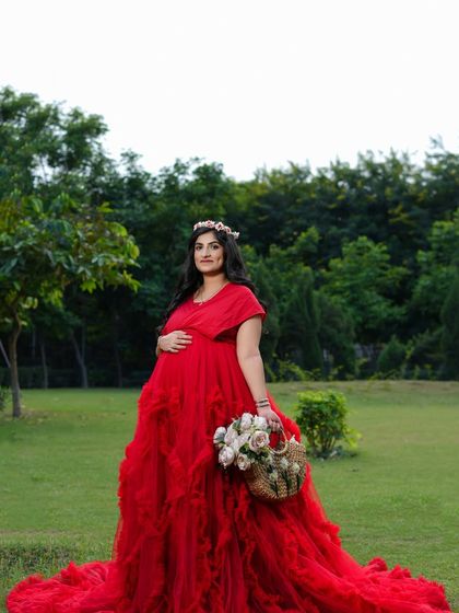 A regal outdoor portrait. The mother-to-be stands tall and serene in a magnificent red gown, holding a basket of flowers like a queen in her garden.