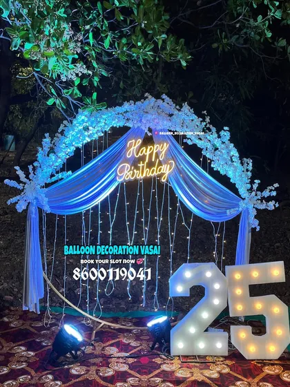 An outdoor birthday setup for a 25th celebration at night. The arch is decorated with white flowers and blue fabric, all illuminated with fairy lights and a "Happy Birthday" neon sign.