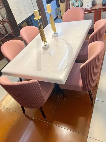 An elegant dining set featuring a white onyx table and chairs upholstered in a pleated pink velvet. The gold-finished table base and chair legs add a touch of warmth and glamour.