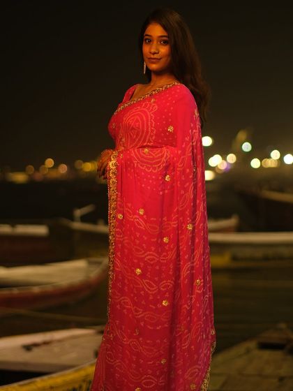 A full-length portrait in a pink saree against the backdrop of boats on the Ganga at night, capturing the festive atmosphere of Dev Deepawali.