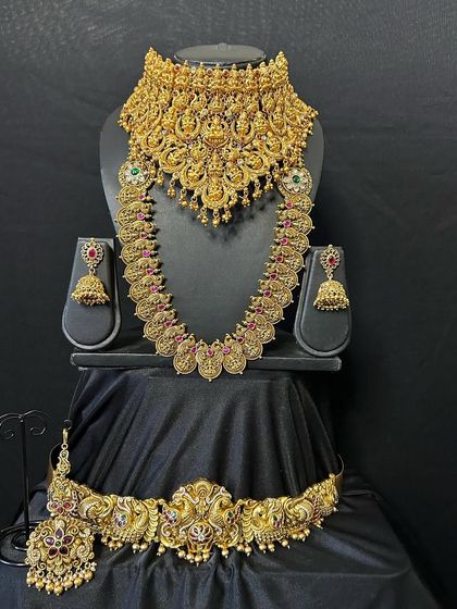 This grand bridal set features a heavy choker and a matching long necklace with goddess Lakshmi motifs. The antique gold finish and red kemp stones give it a very traditional and auspicious feel for a wedding ceremony.